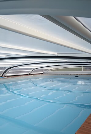 En indendørs swimmingpool med krystalklart vand, omgivet af en moderne arkitektonisk bygning med bløde, kurvede linjer og en træterrasse.