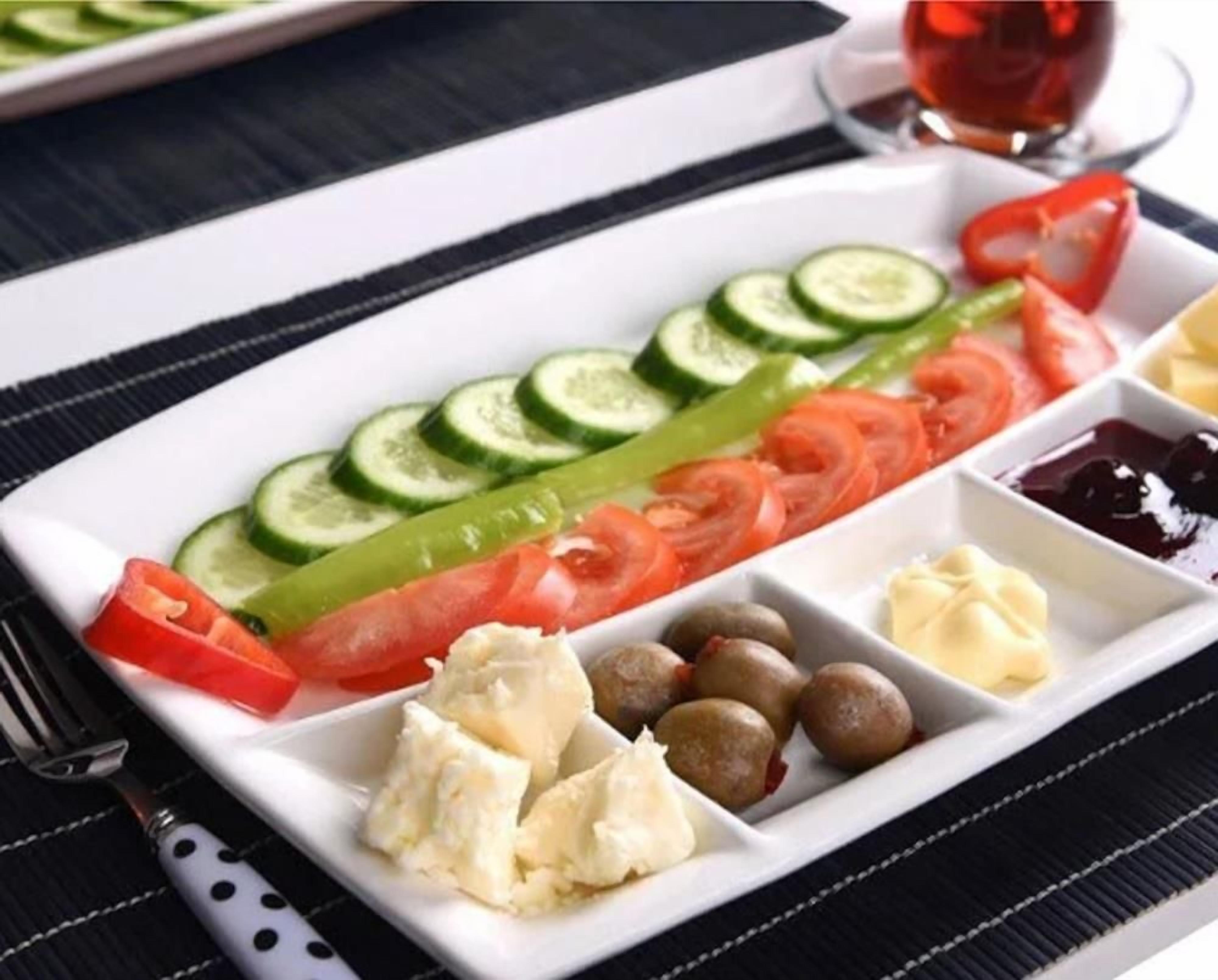 Prato branco retangular com fatias de pepino, tomate, pimentão verde e azeitonas, acompanhado de um garfo e um copo de bebida vermelha.