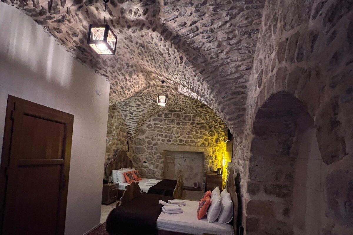 Una habitación con paredes de piedra y techo abovedado, que incluye una cama con almohadas, una mesita y una alfombra.
