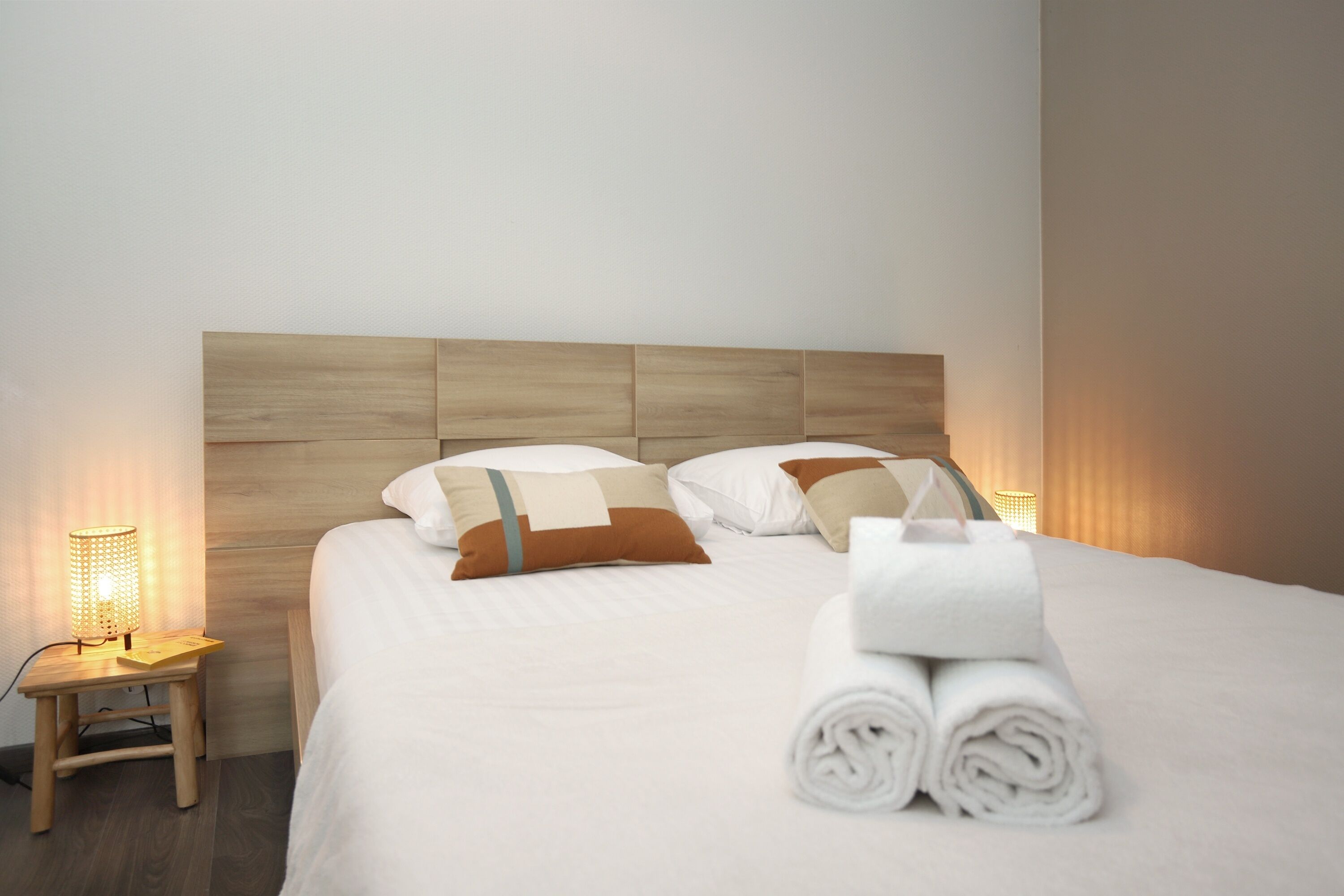 A neatly made bed with a wooden headboard, a small bedside table with a lamp, and rolled towels on the bed.