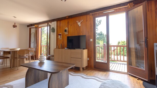 Ein großzügiger Wohnbereich mit Holzboden, einem Esstisch mit Stühlen, einer Couch, einem Fernseher auf einem Holzschrank und einer großen Schiebeglastür, die zu einer Terrasse im Freien führt.