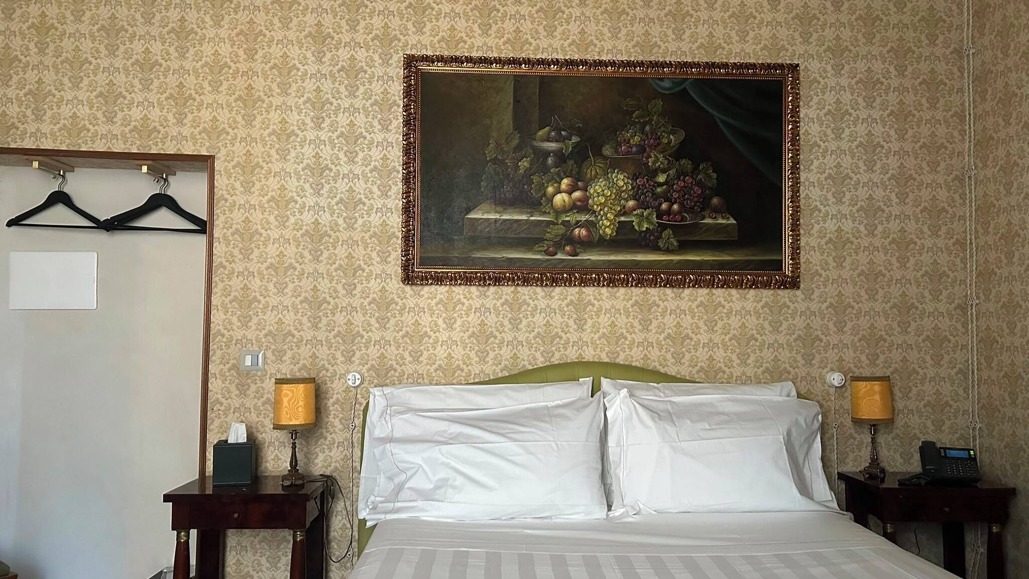 A hotel room with a bed, two bedside tables, a painting of fruit, a coat rack, and a door.