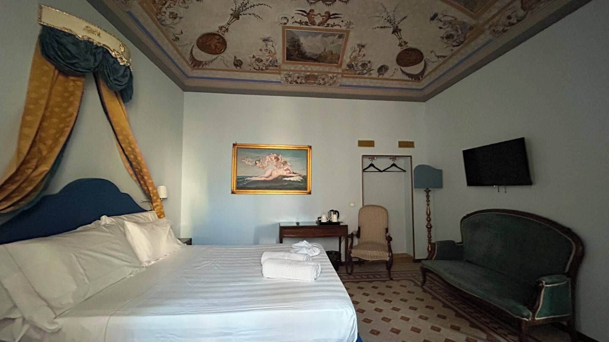 A hotel room with a bed, a chair, a desk, a television, and a painting on the wall.