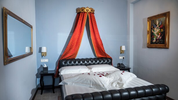 A room with a bed, a nightstand, a lamp, a mirror, and a painting on the wall. - Garden Palace Hotel (Castiglione del Lago)