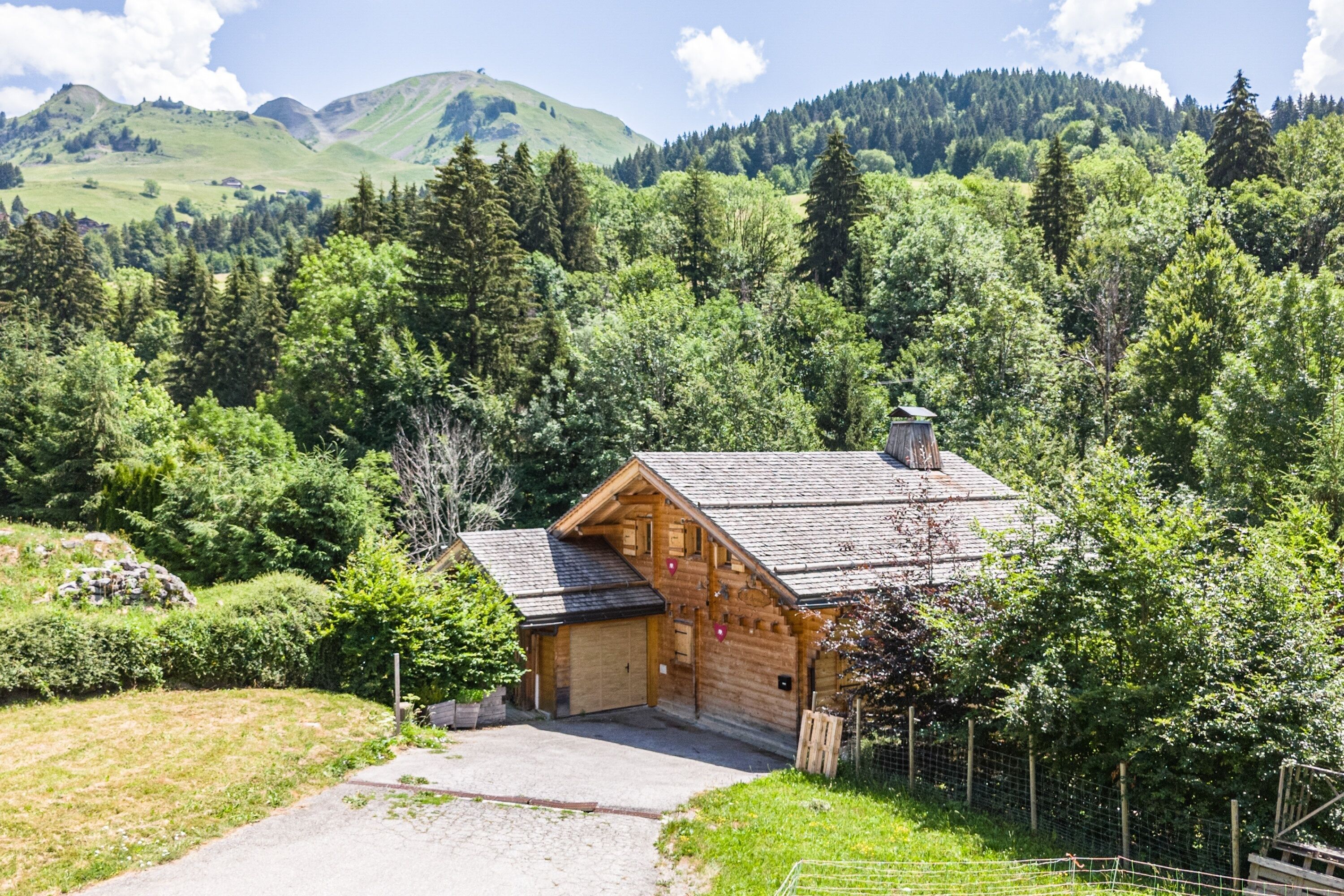 A wooden cabin with a sloped roof, surrounded by dense forest and mountains in the background.