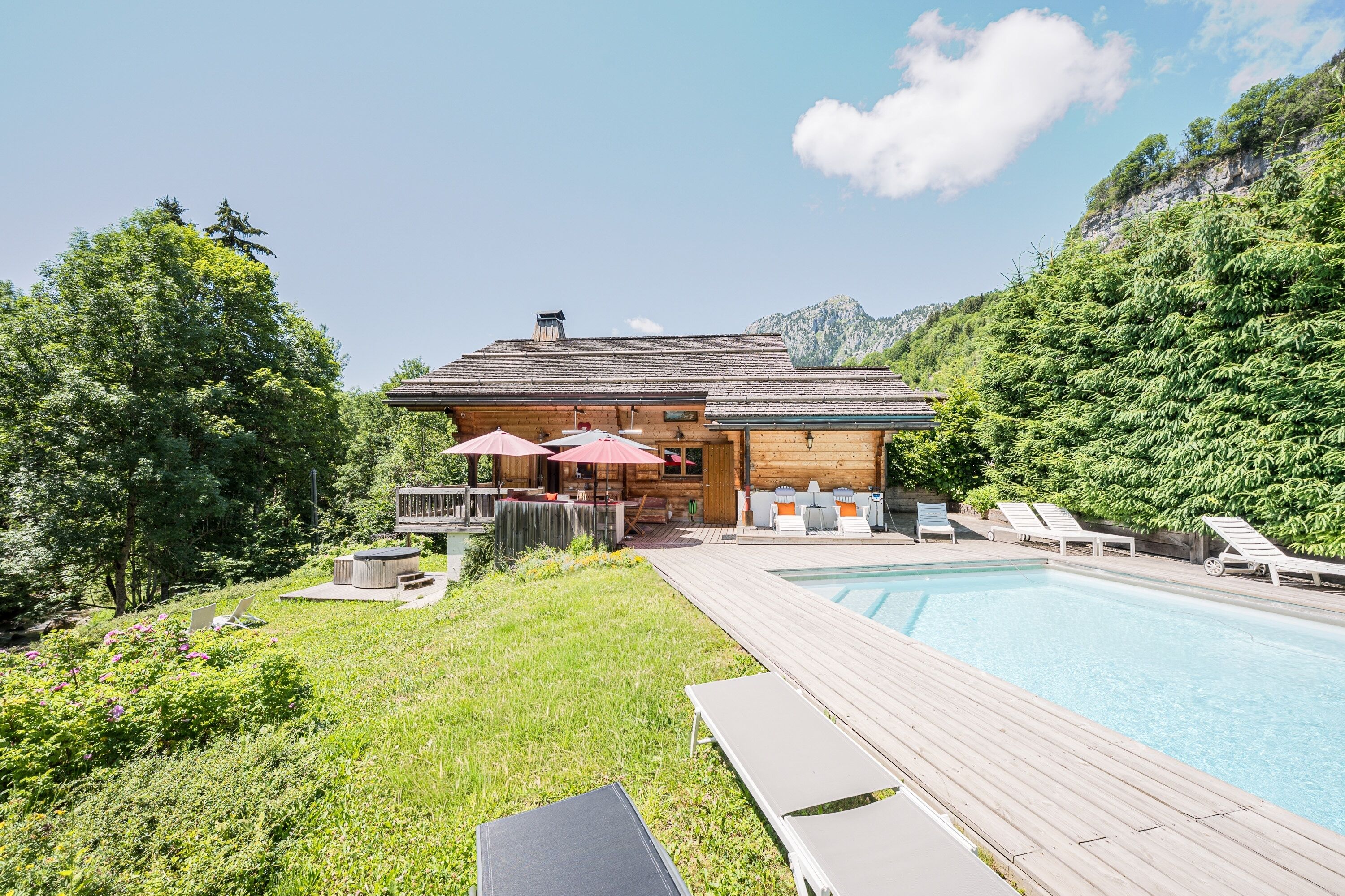 A wooden cabin with a pool, surrounded by greenery and mountains.