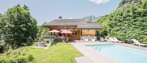 A wooden cabin with a pool, surrounded by greenery and mountains.