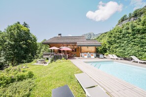 En stuga av trä med en pool, omgiven av grönska och berg.