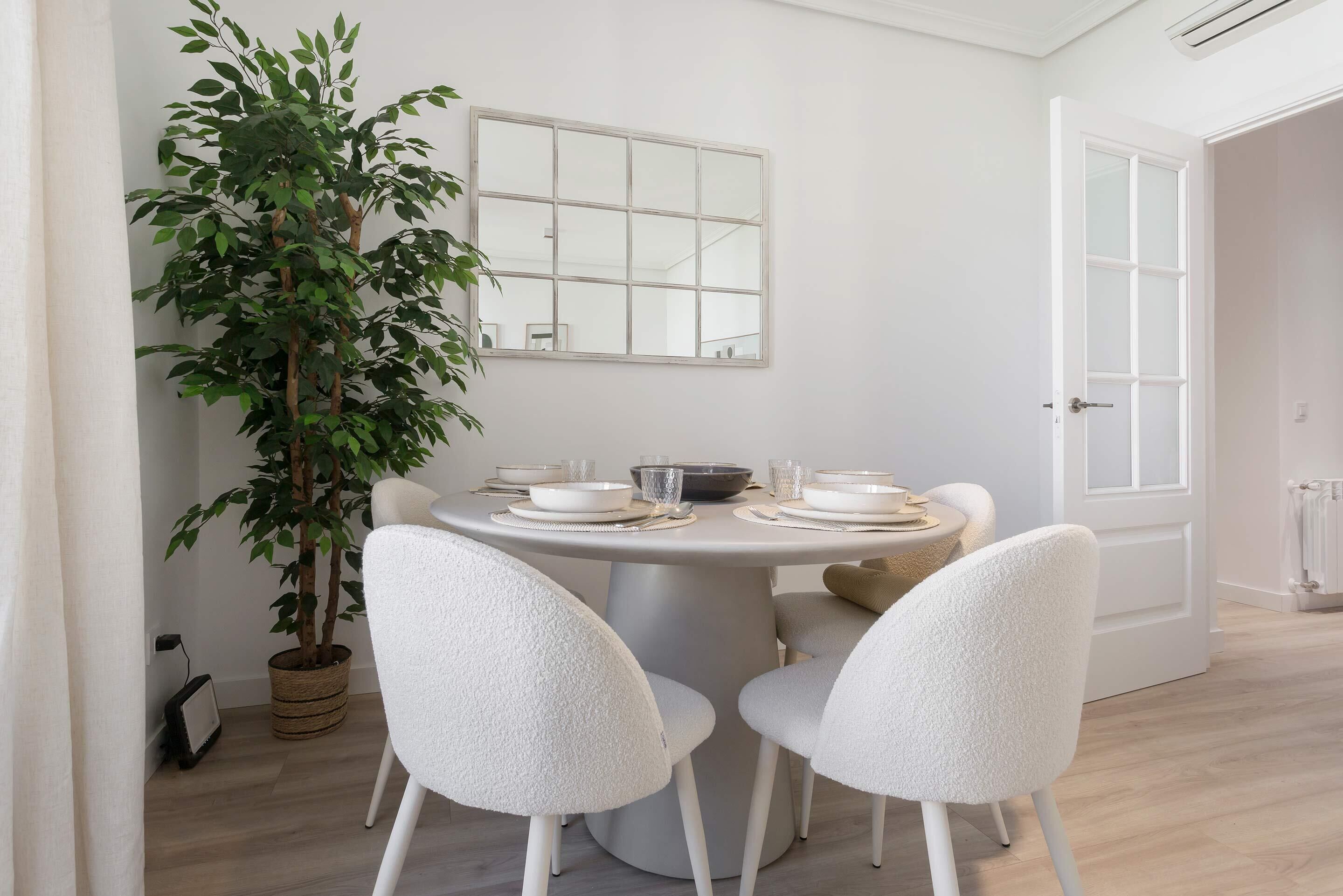 A dining area with a round table set for four, surrounded by white chairs, a potted plant, and a window with a view.