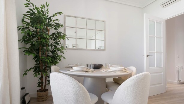 A dining area with a round table set for four, surrounded by white chairs, a potted plant, and a window with a view. - Genteel Home Reina Sofia IV (Madrid)