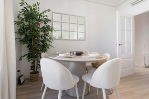 A dining area with a round table set for four, surrounded by white chairs, a potted plant, and a window with a view. - Genteel Home Reina Sofia IV (Madrid)