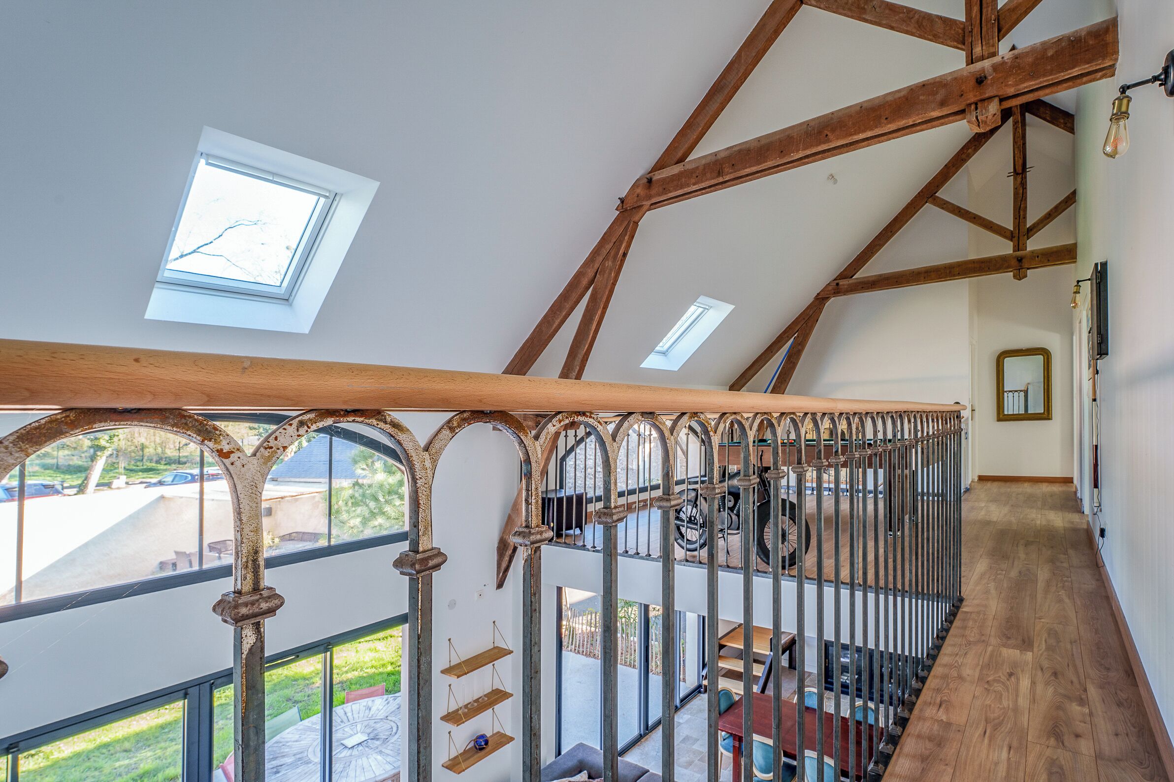 A high-ceilinged room with exposed wooden beams, a staircase with a metal railing, and a view of the outdoors through large windows.