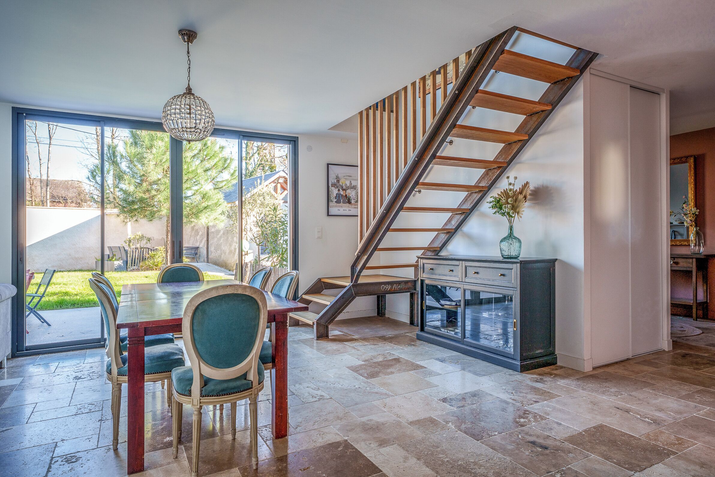 Un vaste espace à manger avec une porte vitrée donnant sur un jardin, un escalier avec des marches en bois et une cuisine moderne équipée d’un four encastré.