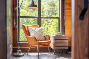 A room with a large window, a leather chair, a wooden cabinet, and a floor lamp. - Cloud Haven Retreat | Views, Hot Tub + Games (Gatlinburg)