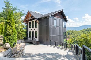 A modern two-story building with a stone foundation, large windows, and a balcony overlooking a forested area. - Cloud Haven Retreat | Views, Hot Tub + Games (Gatlinburg)