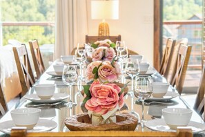 Ein Esszimmertisch ist für eine Mahlzeit gedeckt, mit Stühlen, Gläsern und einer Blumenarragement mit rosa Blumen.