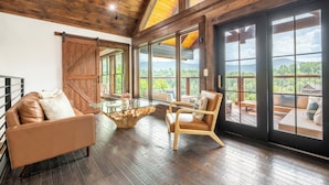 A spacious room with a large sliding barn door, a glass coffee table, leather sofas, and a view of a mountainous landscape. - Cloud Haven Retreat | Views, Hot Tub + Games (Gatlinburg)