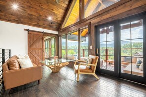 A spacious room with a large sliding barn door, a glass coffee table, leather sofas, and a view of a mountainous landscape. - Cloud Haven Retreat | Views, Hot Tub + Games (Gatlinburg)