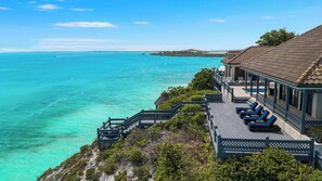 A coastal property with a deck overlooking turquoise waters, a covered patio with lounge chairs, and a thatched-roof structure.