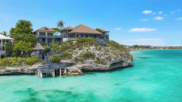 A beachfront property with multiple buildings, a staircase leading to a deck, and clear turquoise waters.