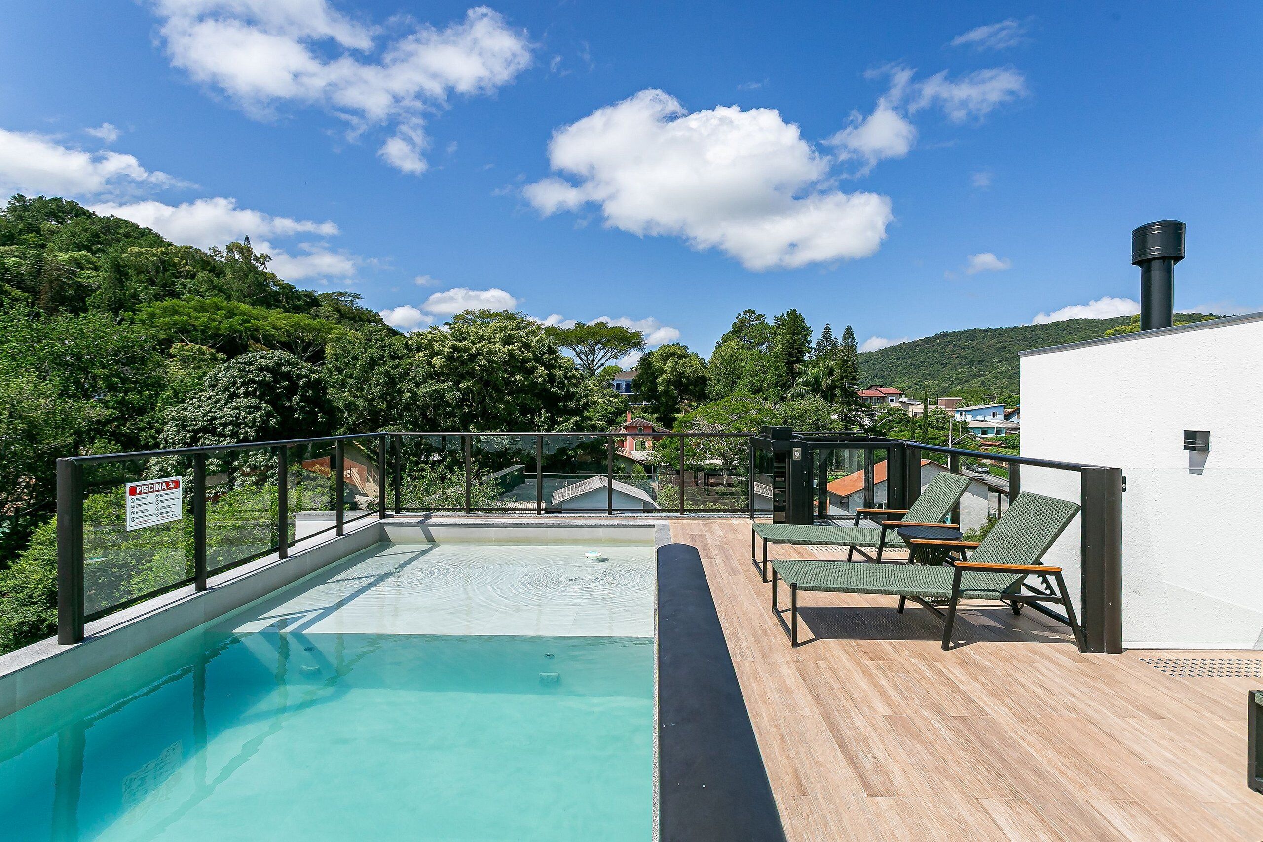 Piscina com deck de madeira, espreguiçadeiras e vista para uma colina com árvores e casas.