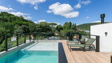 Piscina com deck de madeira, espreguiçadeiras e vista para uma colina com árvores e casas.