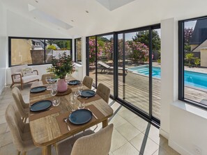A dining area with a table set for six, large windows, and a view of a swimming pool. - House for 6, pool, boules & foosball (Plomeur)