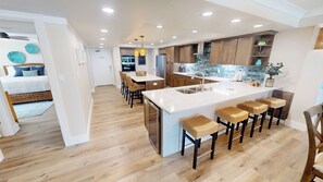 A modern kitchen with a central island, bar stools, wooden cabinets, and a dining area with wicker chairs. - Stunning & Renovated 2 Bedroom ST 2105 (Destin)