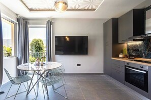 Une cuisine moderne équipée d’un téléviseur à écran plat, d’un coin repas avec une table ronde et des chaises, et d’un balcon offrant une vue sur la verdure.