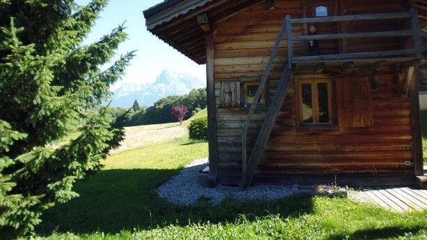 En træhytte med en trætrappe op til en balkon, omgivet af grøn bevoksning og bjerge i baggrunden.