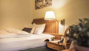 Een hotelkamer met een opgemaakt bed, een bedlampje, een nachtkastje met een telefoon en een tijdschrift, en een kunstwerk aan de muur.