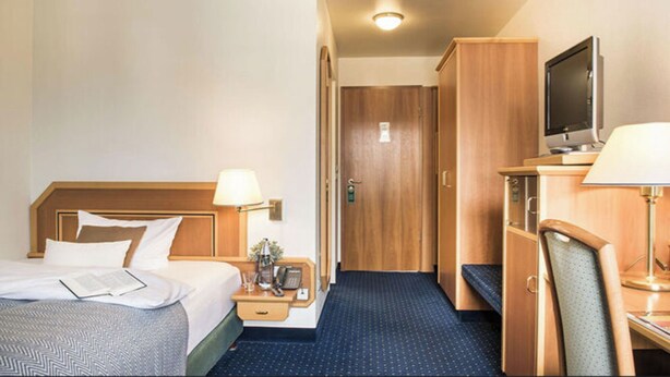 A hotel room with a bed, bedside table, lamp, TV, and a wardrobe. - Explore Lakes & Forests | Single Room | Sauna & Dining Experience (Sundern (Sauerland))