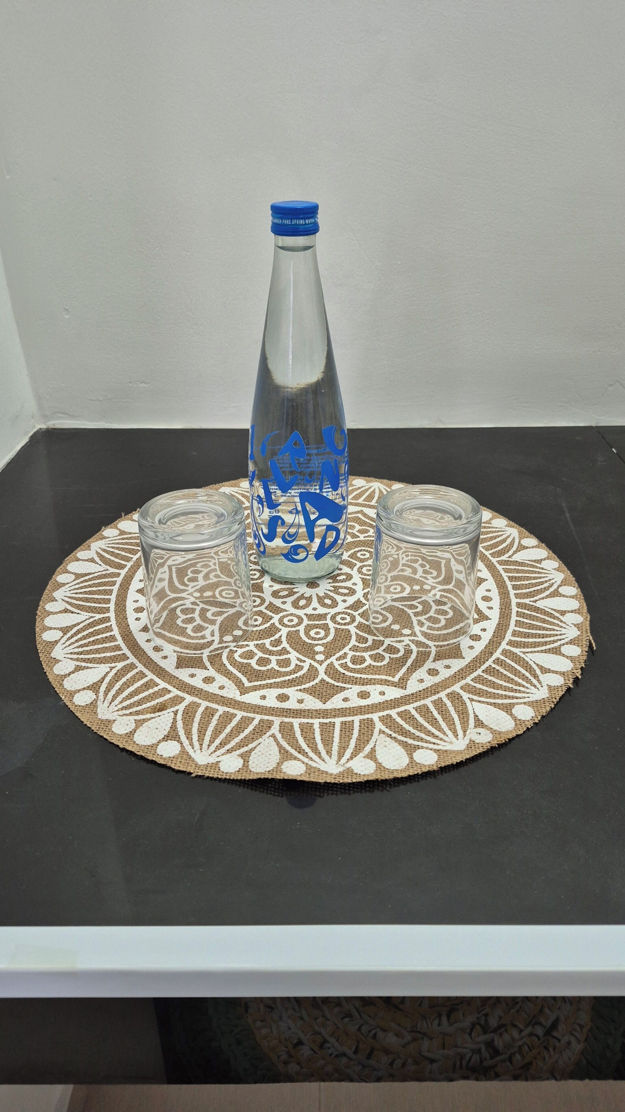 Una botella de vidrio transparente con texto azul sobre una mesa con un tapete decorativo.