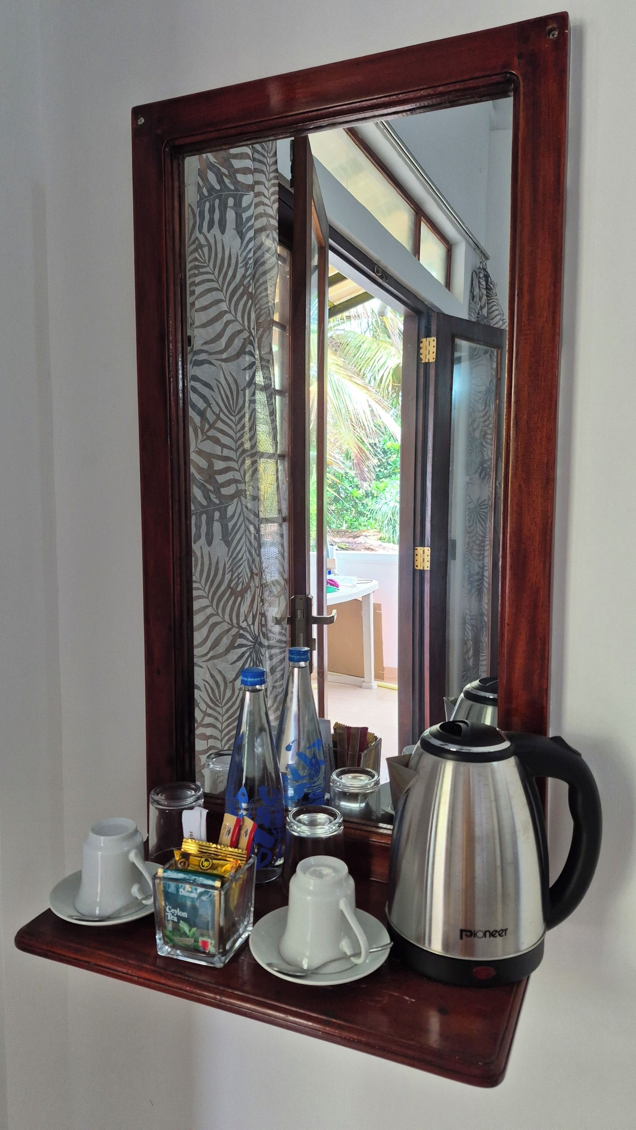 Un espejo refleja una habitación con un estante de madera que contiene una tetera blanca, una pava y varias botellas.