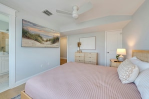 Une chambre à coucher comprenant un lit, un ventilateur de plafond, une commode, une table de chevet avec une lampe et un tableau encadré accroché au mur.