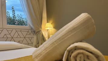 A neatly made bed with a rolled towel on it, a lamp on a bedside table, and a window with curtains.