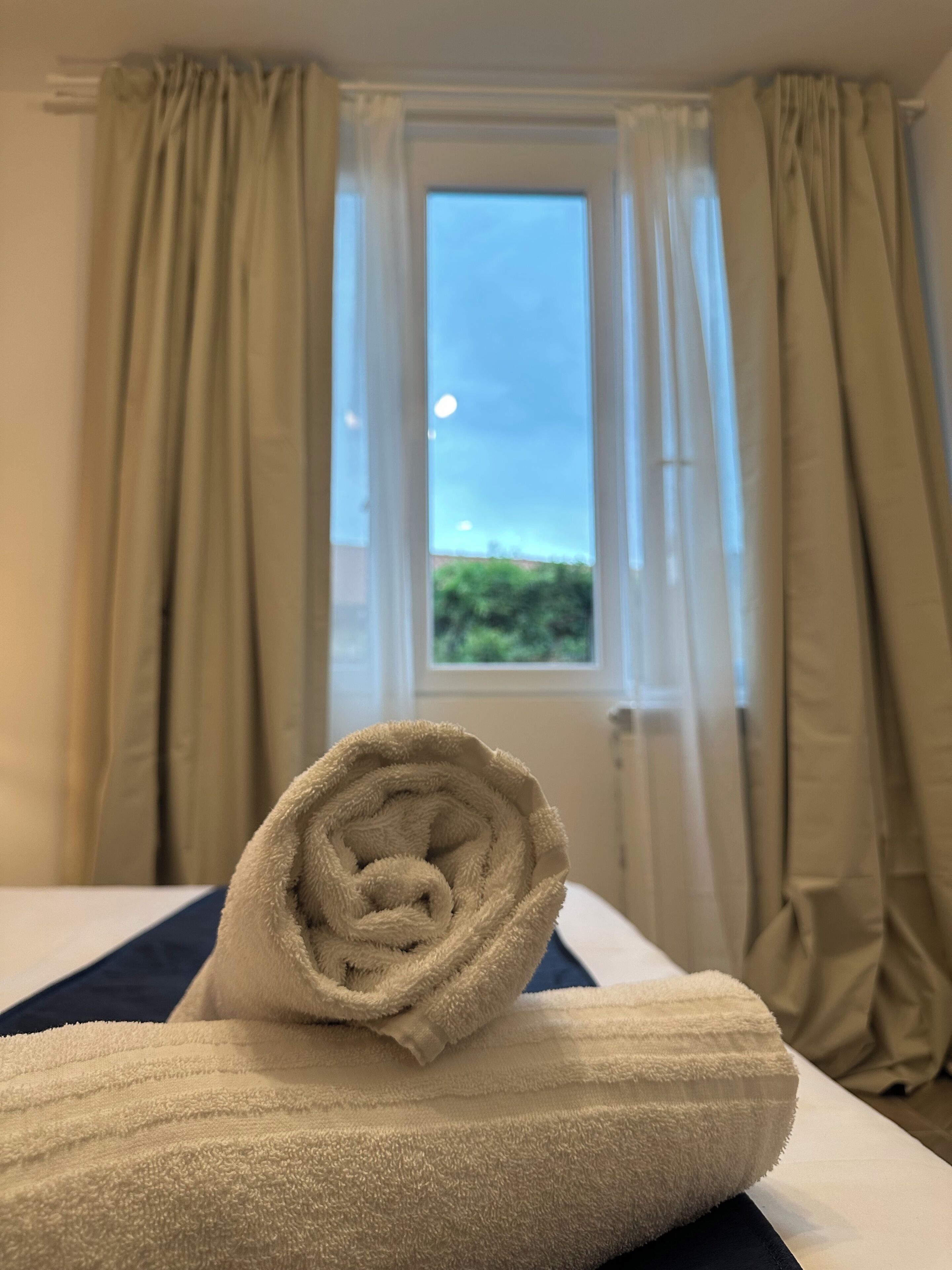 A neatly made bed with a rolled white towel in the center, a window with a view of greenery, and beige curtains.