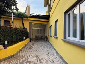 Un edificio amarillo con un paseo empedrado, entrada por puerta de cristal y un pequeño espacio exterior con mesas.
