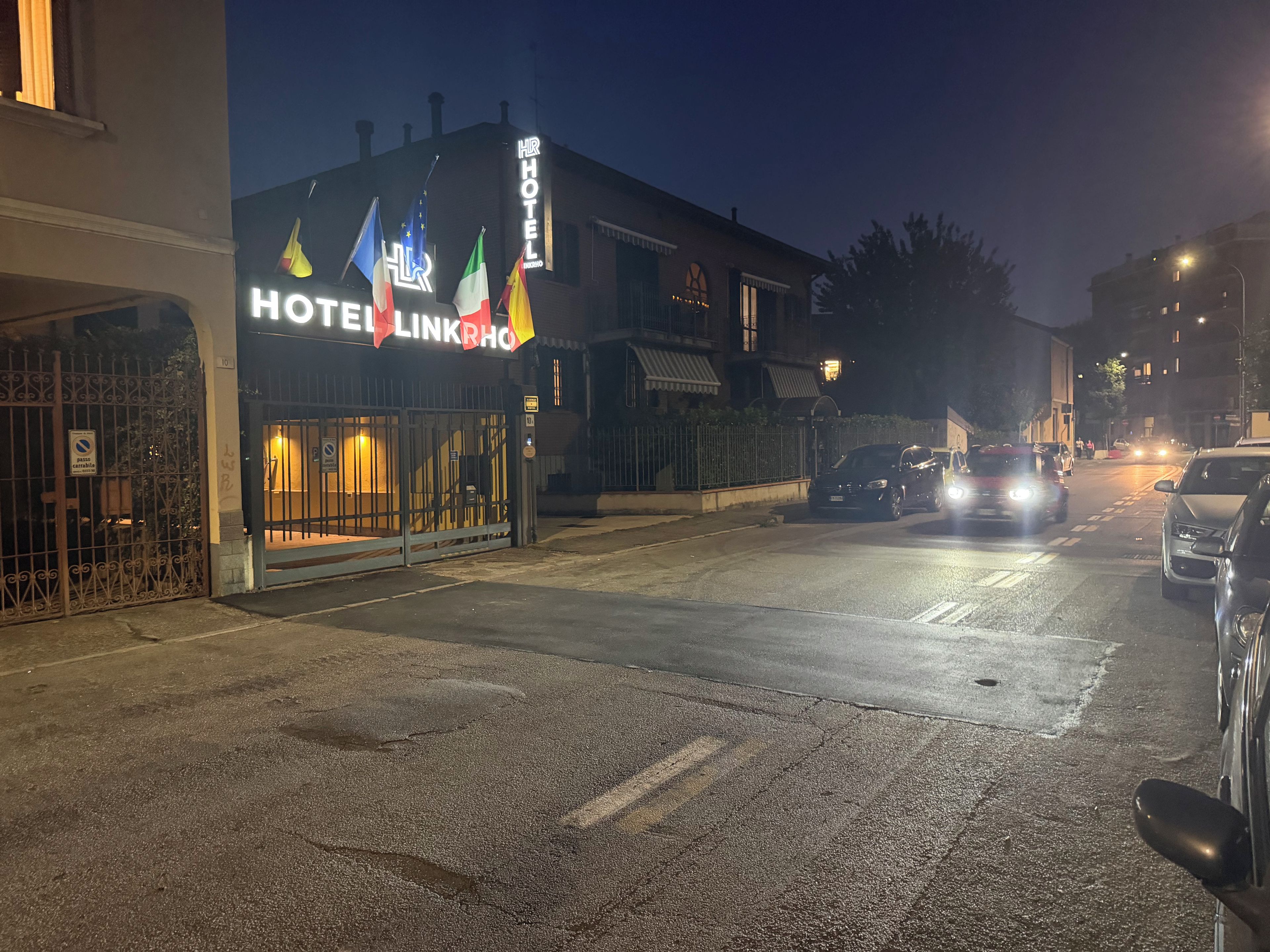 Un hôtel avec une enseigne, une entrée fermée par un portail et une rue bordée de voitures garées.