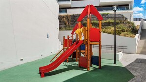 A playground with a red slide and climbing structures, set against a white wall with a green ground cover. - Rooftop* Terrace*pool*parking* Town view (Estepona)