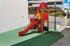 Um parquinho com um escorrega vermelho e estruturas de escalada, em frente a uma parede branca e com cobertura de grama verde.