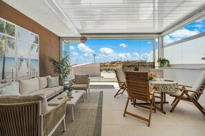 Uma sala de estar moderna com um sofá, cadeiras, uma mesa de jantar e vista para o mar através de janelas amplas.