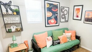 Un salon avec un canapé vert, divers coussins, une petite table avec une lampe, un panier et des œuvres d’art murales comprenant un chat, des disques vinyle et une enseigne « Route 66 ».