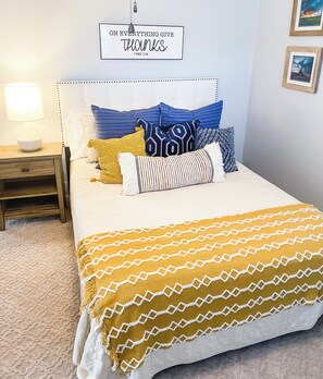 A bed with a headboard, multiple pillows, a bedside table with a lamp, and a wall with a framed quote.
