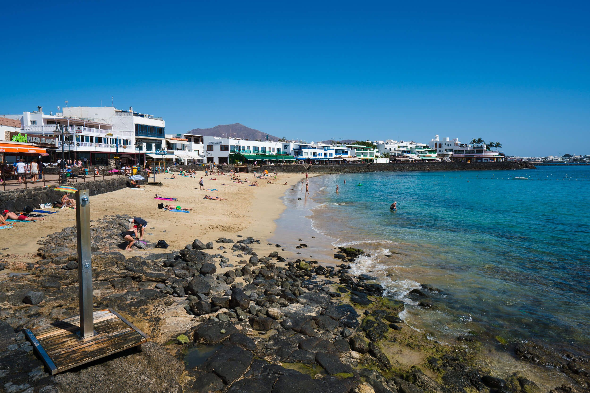 Ein Strand mit felsigem Ufer, klarem, blauem Wasser und Gebäuden im Hintergrund.