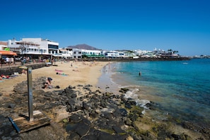 Une plage au rivage rocheux, avec une eau d’un bleu limpide et des bâtiments visibles à l’arrière-plan.