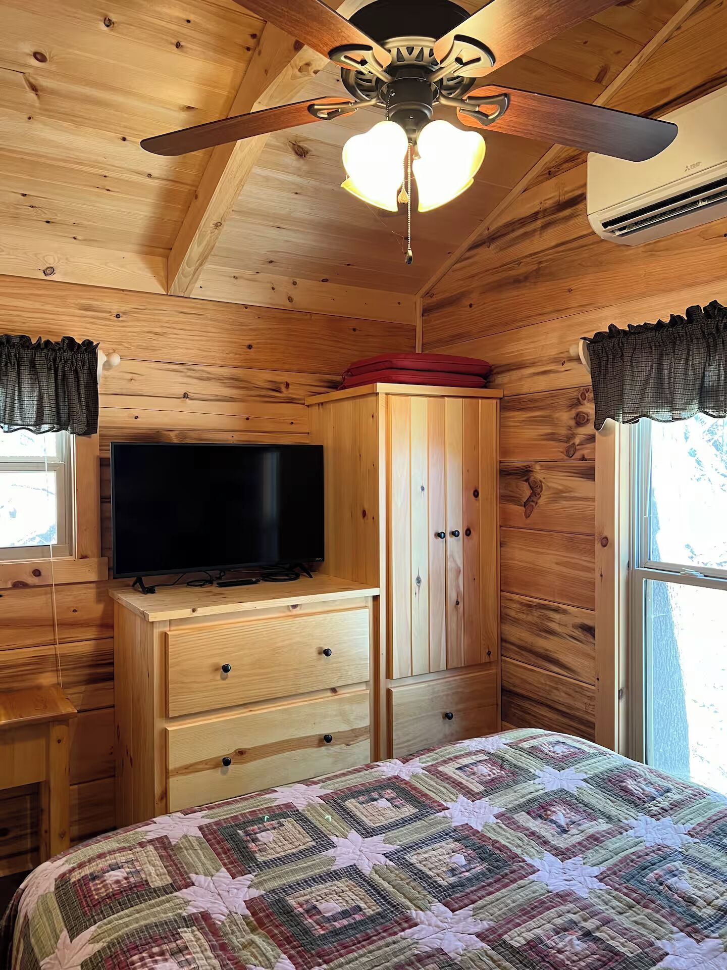 Ein Zimmer einer Holzhütte mit Deckenventilator, einem Fernseher auf einer Holzkommode, einem Kleiderschrank und einem Bett mit Steppdecke.