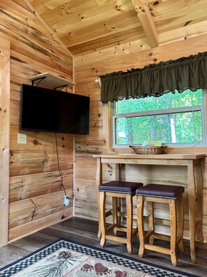 A wooden interior with a television mounted on the wall, a window with a view of greenery, and a small table with a basket.