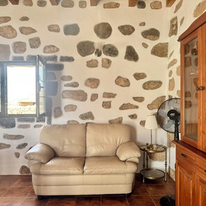 A room with a stone wall, a leather sofa, a wooden cabinet, and a fan. - Casa Clarita (Tindaya)