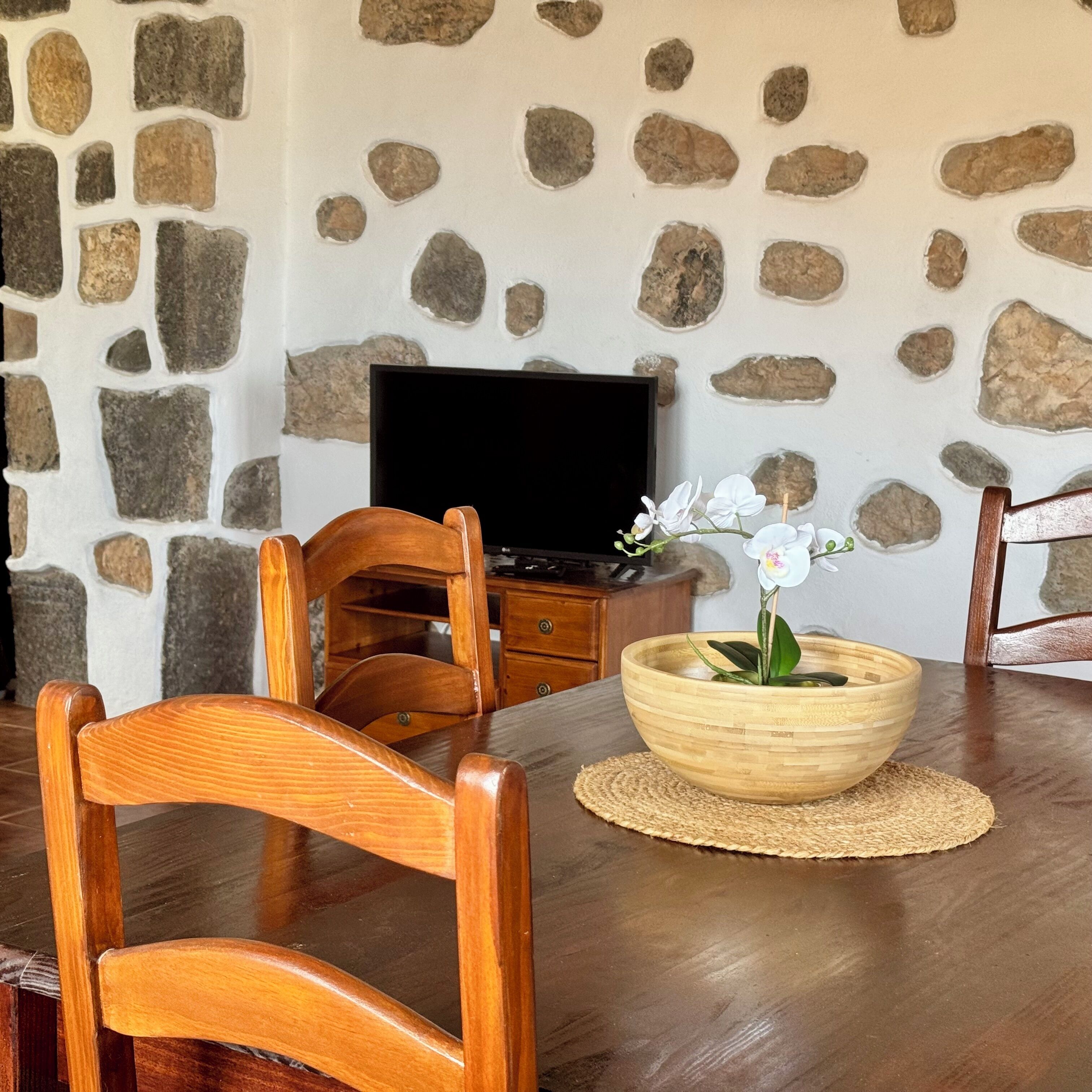 A dining area with a stone wall, wooden chairs, a television, and a bowl with flowers.